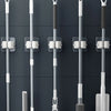 A collection of rod-shaped objects, including mops and brushes, are shown hanging on adhesive wall hooks.