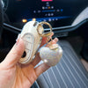 A hand holds a peach heart crystal keychain charm attached to a white bag charm inside a car interior.