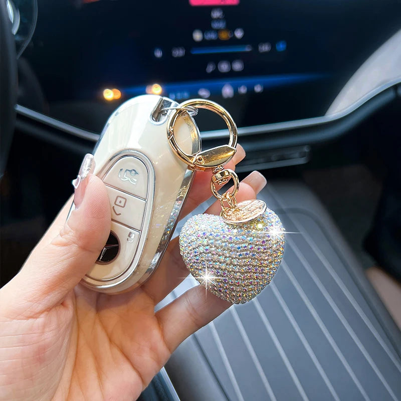 A hand holds a peach heart crystal keychain charm attached to a white bag charm inside a car interior.