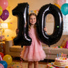 A young girl is holding giant black foil balloons shaped like the number 10 in a festive indoor setting.