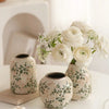A white ceramic vase with green floral patterns holds a bouquet of white roses and baby's breath flowers.