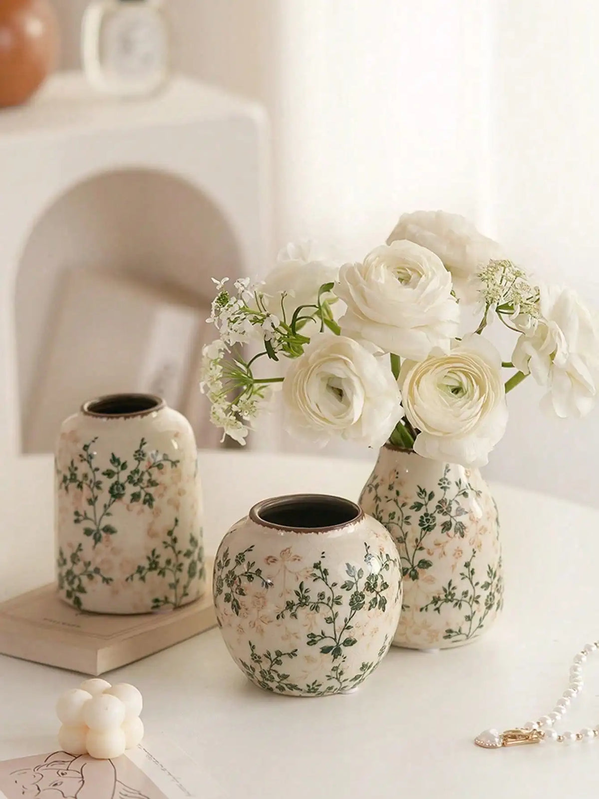 A white ceramic vase with green floral patterns holds a bouquet of white roses and baby's breath flowers.