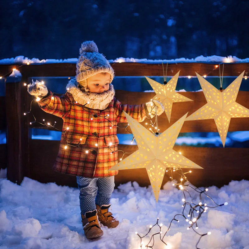 A child wearing a knitted hat and plaid coat holds up several glowing star-shaped pendant lights in a snowy outdoor setting.
