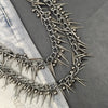A close-up of a silver metal chain necklace featuring sharp rivets and spikes against a gray fabric background.