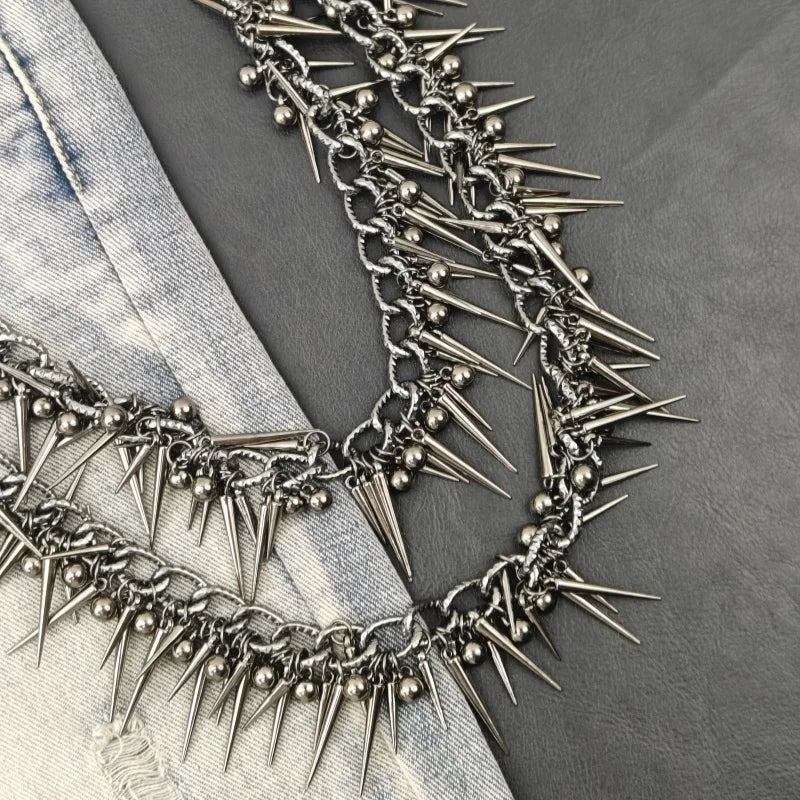 A close-up of a silver metal chain necklace featuring sharp rivets and spikes against a gray fabric background.
