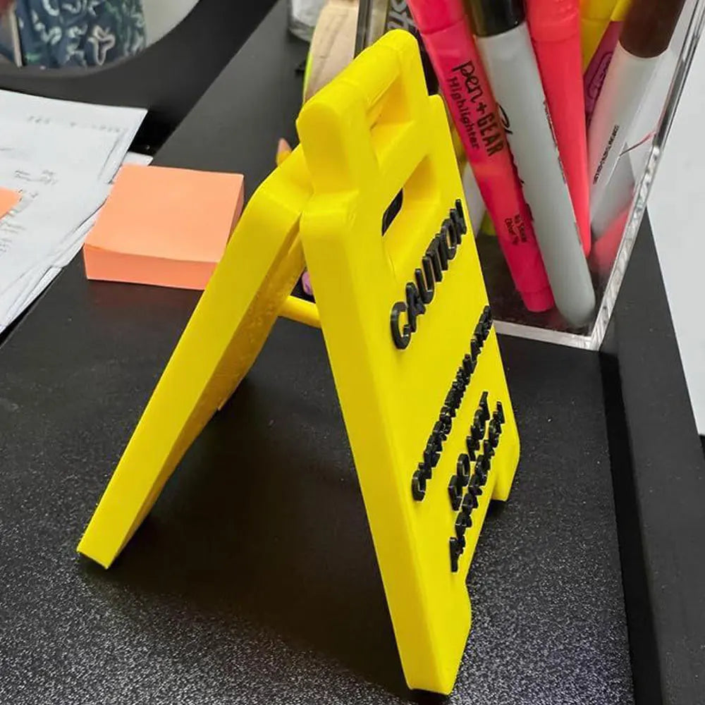 A bright yellow plastic desk sign with black lettering is shown on a dark surface.
