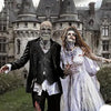 A person wearing a realistic latex skull mask and dark clothing stands in front of a gothic castle backdrop.