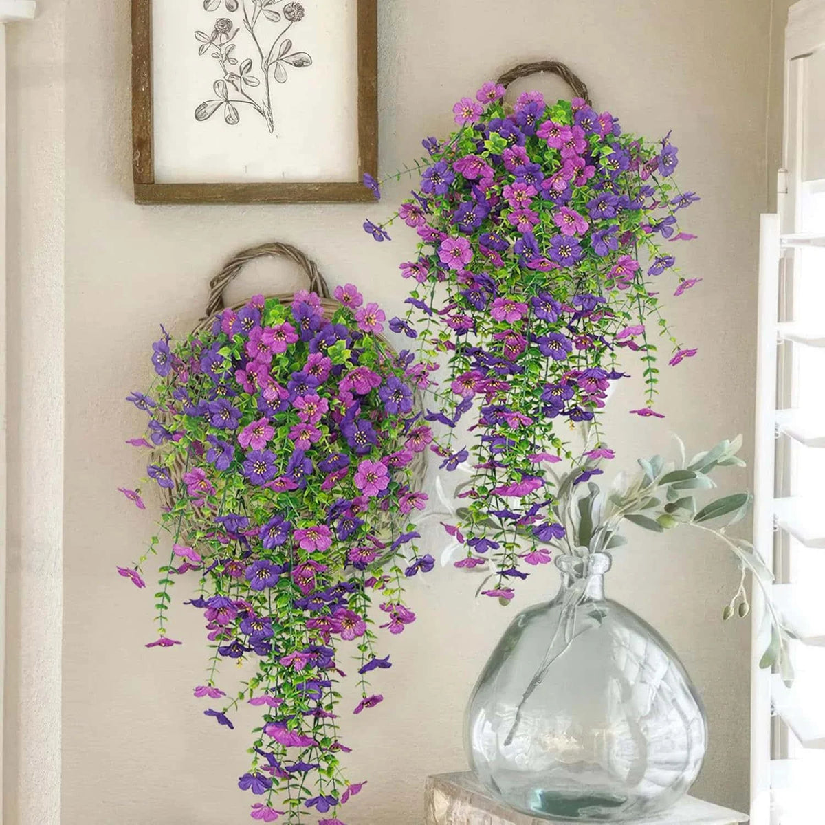A glass vase filled with purple and green faux silk flowers displayed on a light-colored wall.