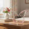 An elegant pink acrylic butterfly plaque with floral designs sits on a wooden surface next to a white vase of roses.