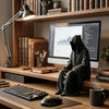 A dark hooded figure sits next to a laptop on a wooden desk with books and office supplies in the background.