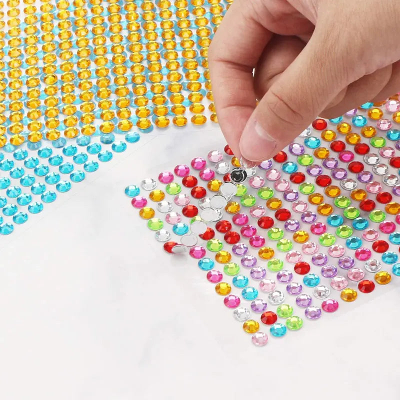 A hand is placing colorful self-adhesive rhinestones onto a white surface.