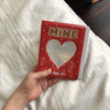 A red photo album with a heart cutout and the text MINE I LIKE IT is held in a hand on white bedding.