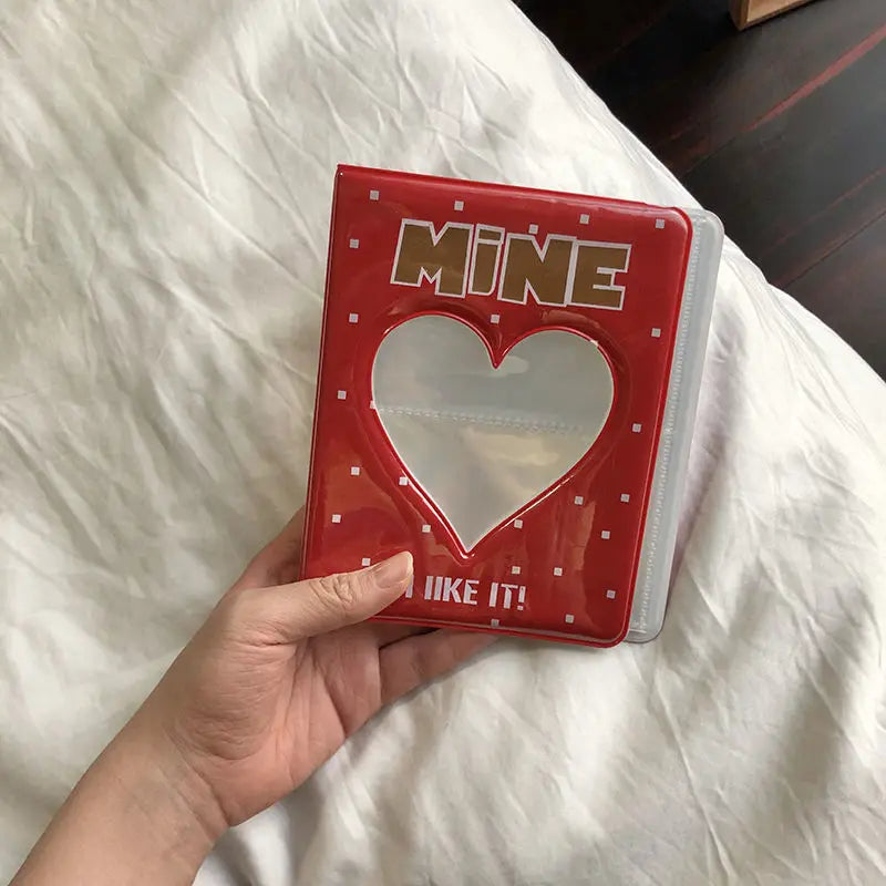 A red photo album with a heart cutout and the text MINE I LIKE IT is held in a hand on white bedding.