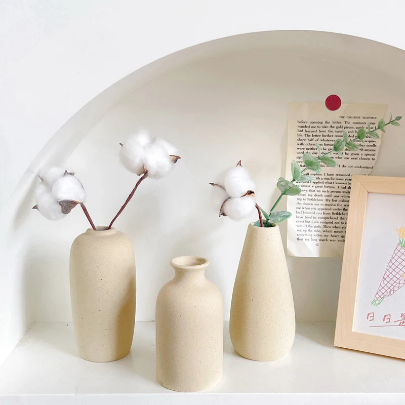 Three minimalist ceramic vases in beige tones displayed on a white shelf with a framed card.
