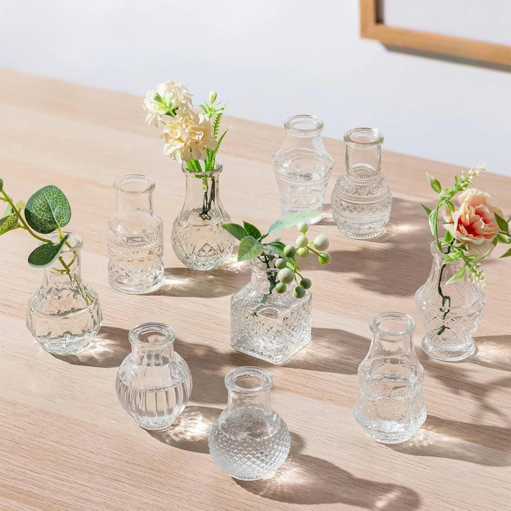 A collection of clear diamond faceted glass bud vases holding small flowers and greenery on a wooden surface.