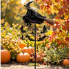 A whimsical witch wind rotor garden decor featuring a black witch hat and feathered wings standing on a metal stake amidst au