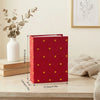 A red laminated photo album with gold heart accents sits on a wooden surface next to a white vase and dried greenery.