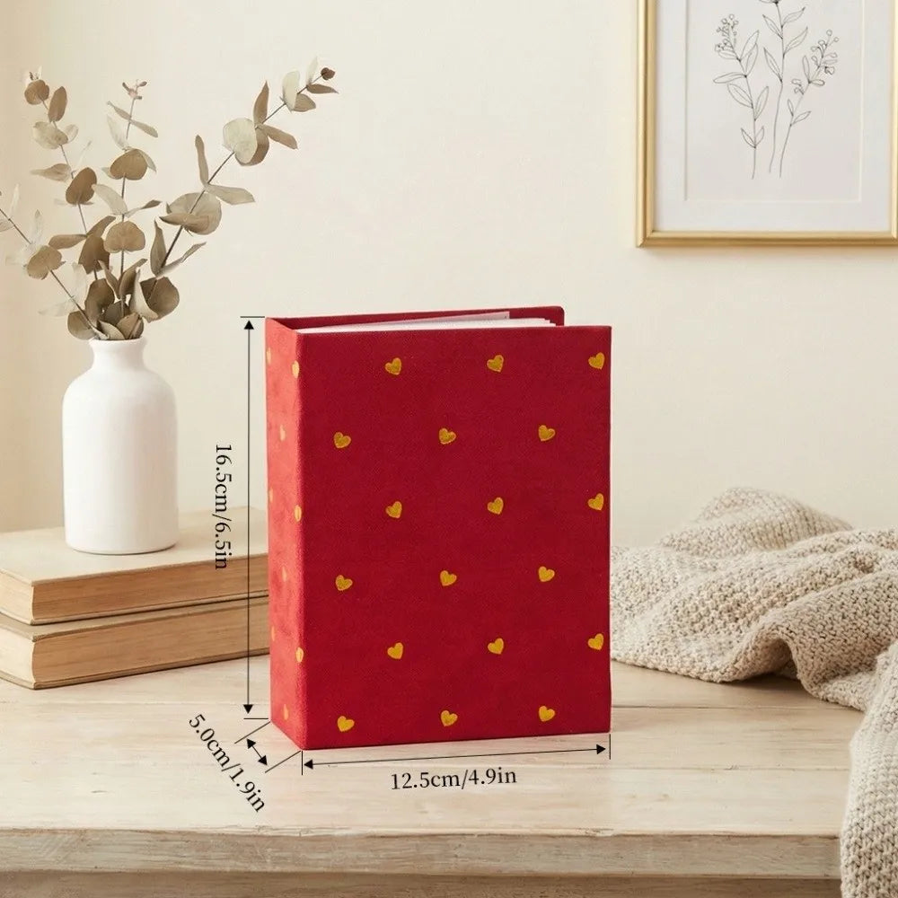 A red laminated photo album with gold heart accents sits on a wooden surface next to a white vase and dried greenery.