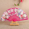 Pink floral silk folding fan displayed on a wooden stand with pink flowers hanging above.
