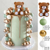 A tiered cake topper featuring a brown teddy bear and surrounding pink, gold, and mint green ball decorations.