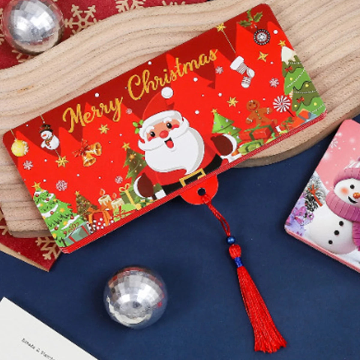 A festive red Christmas card with Santa Claus and gifts, lying on a dark blue surface with a silver ornament.