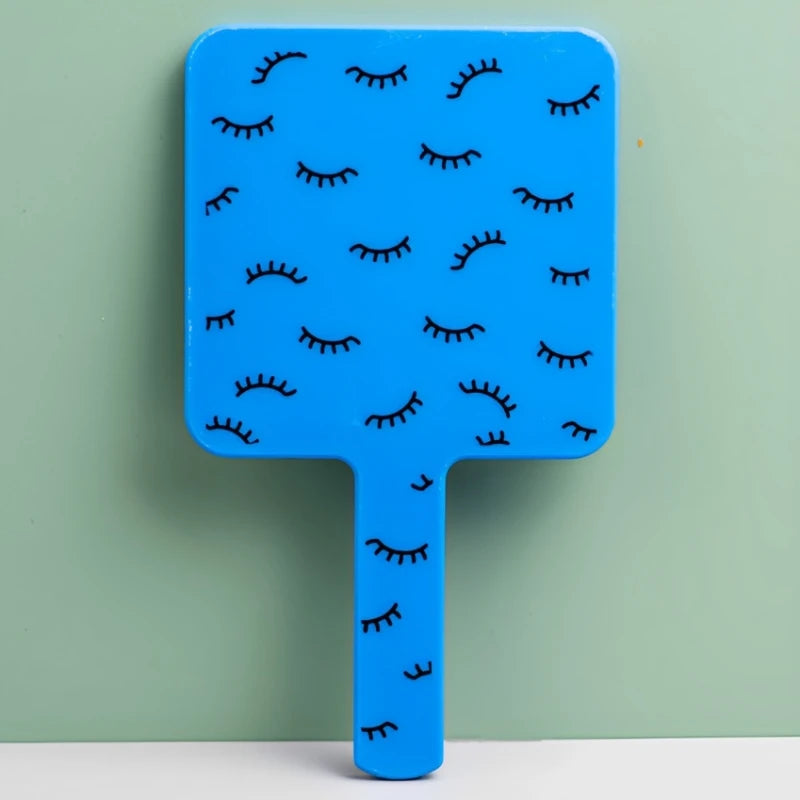 A bright blue handheld vanity mirror with an ergonomic handle featuring a pattern of black eyelashes.