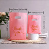 Pink paper gift bags with thank you message and handles displayed on a wooden surface.