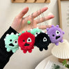 A hand holds several colorful plush keychains with clear photocard holders, featuring green, red, black, and purple designs.