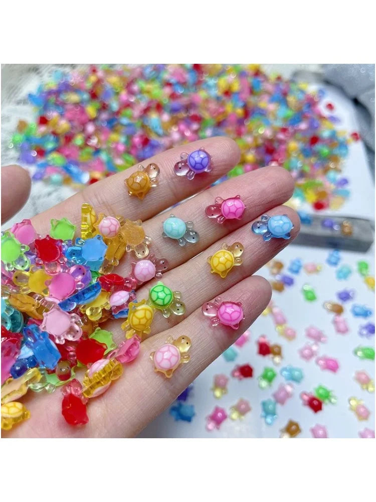 Una mano sostiene una pila de 50 adornos/charms de uñas de tortugas mini y coloridos sobre una superficie blanca.