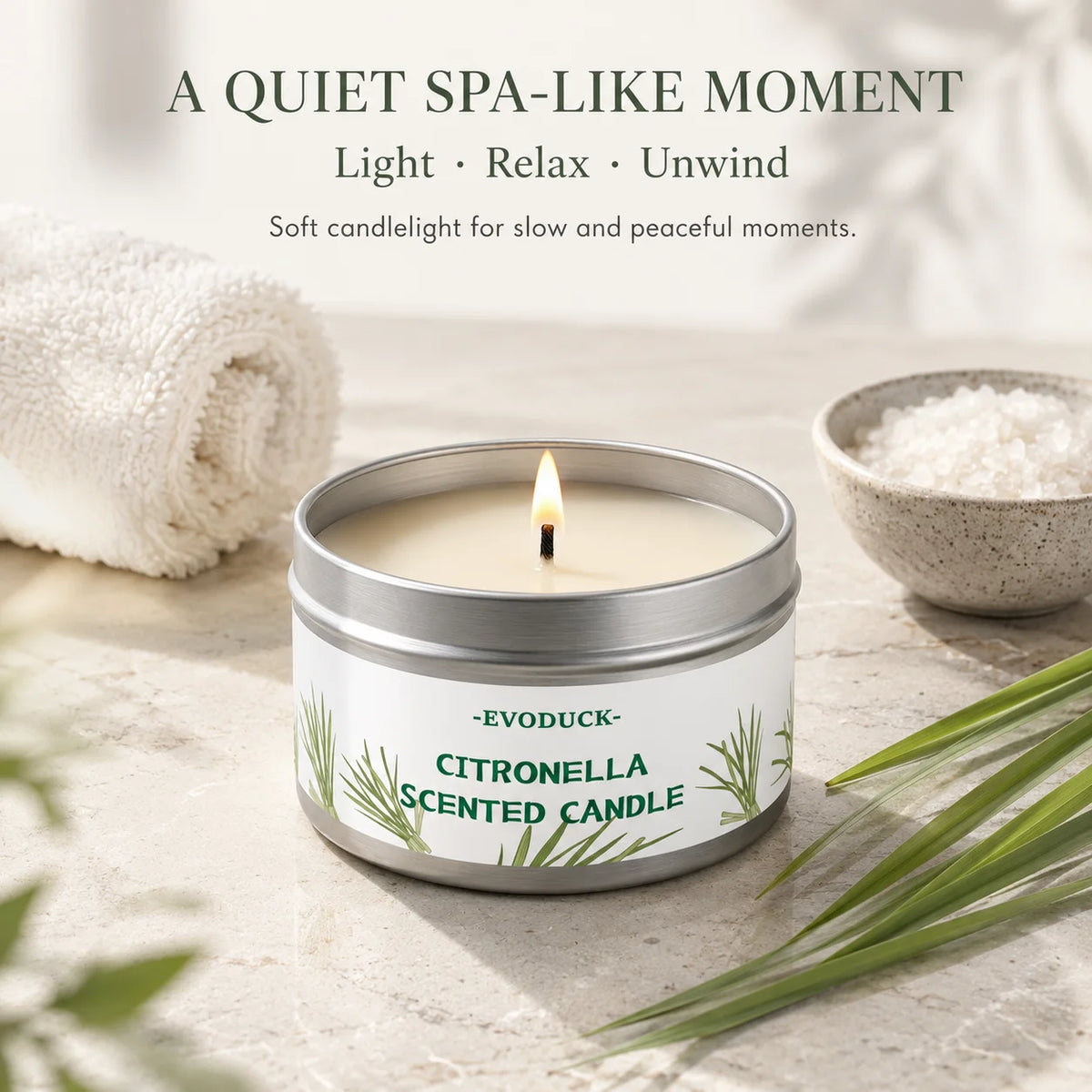 A jar of citronella scented candle sits on a light surface with a white towel and green leaves.