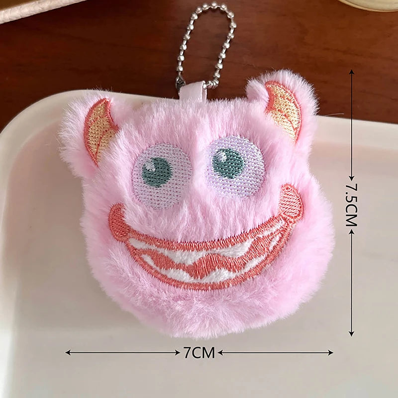 A pink fuzzy plush monster keychain with large eyes and a wide smile is displayed on a white surface.