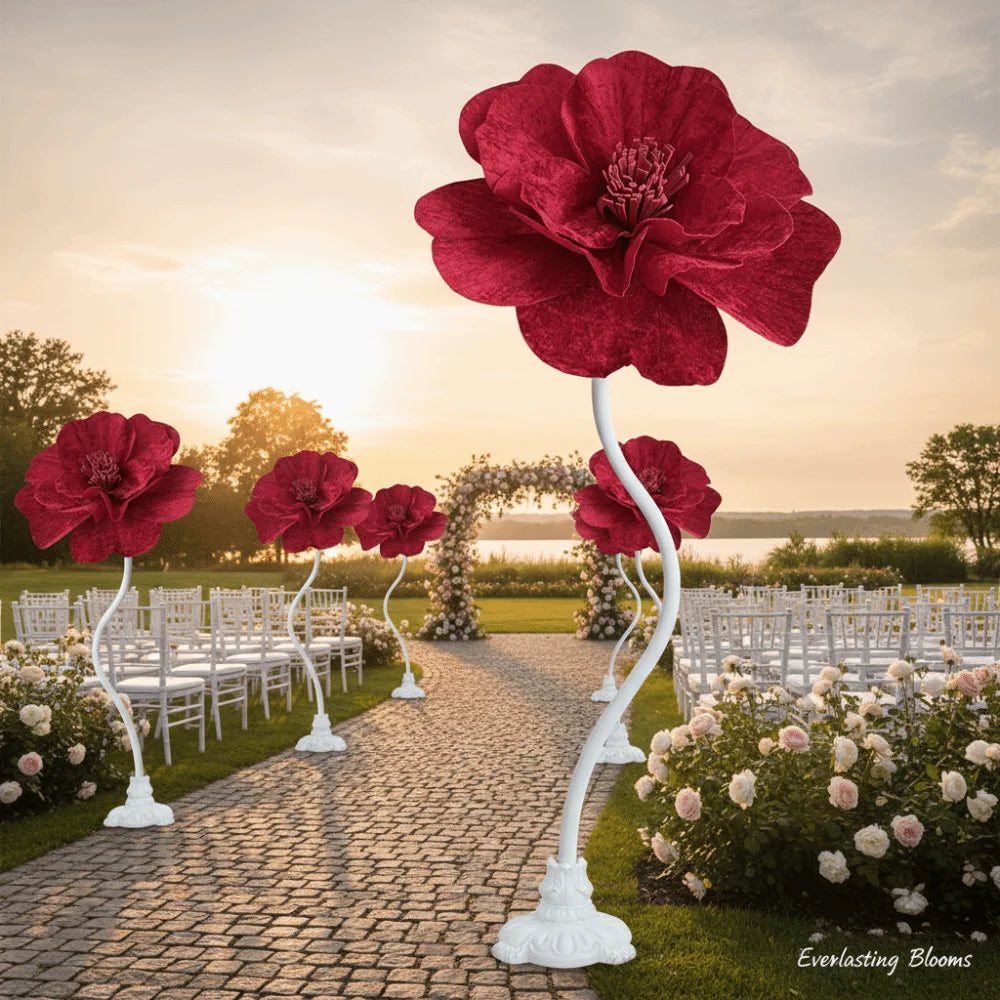 A display of large, deep red velvet artificial peony flowers arranged on white stands in a garden setting.