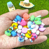 A hand holds a collection of mini resin cruise ducks with sunglasses in assorted colors on green grass.