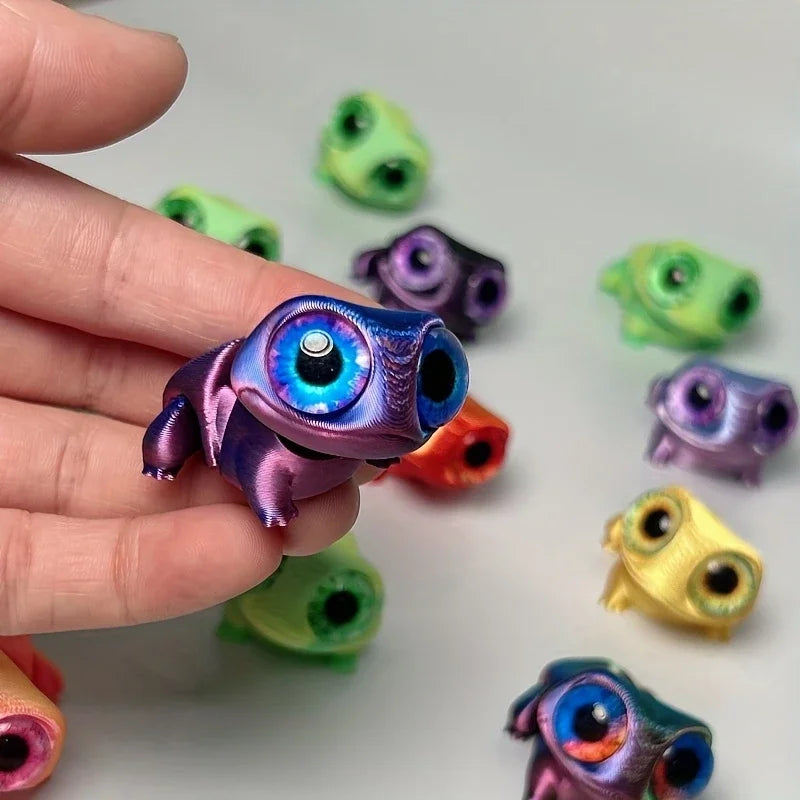 A hand holds a 3D printed frog car ornament with color-shifting eyes, featuring various shades of green and purple.