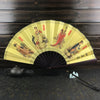 A yellow decorative folding fan with traditional Asian scenes and red accents displayed on a dark wooden surface.