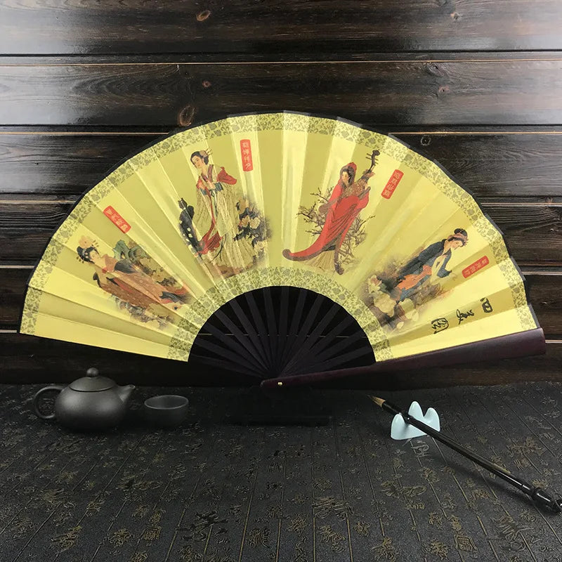 A yellow decorative folding fan with traditional Asian scenes and red accents displayed on a dark wooden surface.
