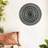 Decoración de pared en forma de mandala de madera negra montada en una pared blanca junto a un cojín de terracota y vegetació