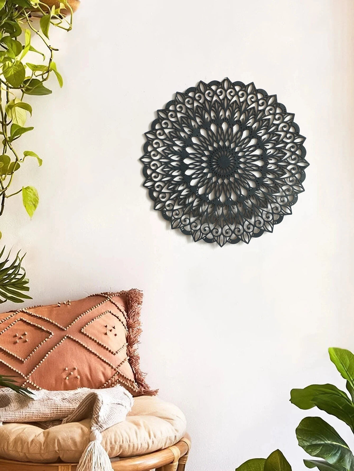Decoración de pared en forma de mandala de madera negra montada en una pared blanca junto a un cojín de terracota y vegetació
