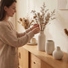 A woman places dried floral branches into a set of three matte, vertically ribbed ceramic vases on a wooden cabinet top.