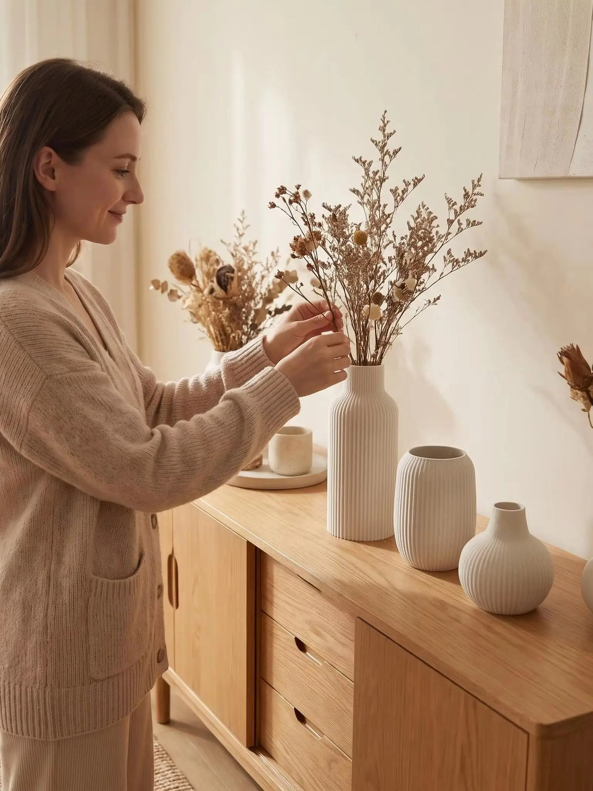 A woman places dried floral branches into a set of three matte, vertically ribbed ceramic vases on a wooden cabinet top.