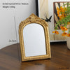 A gold-framed arched carved mirror with a reflective surface sitting on a dark wooden table.