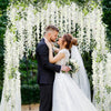 A bride and groom stand under a white floral garland archway in a garden setting.