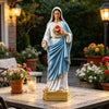 A statue of the Blessed Mother Mary with a white and blue robe and a golden crown, displayed on an outdoor table.