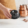 A hand holds a black nonstick mini fluted cake pan above a stack of copper baking pans on a white countertop.