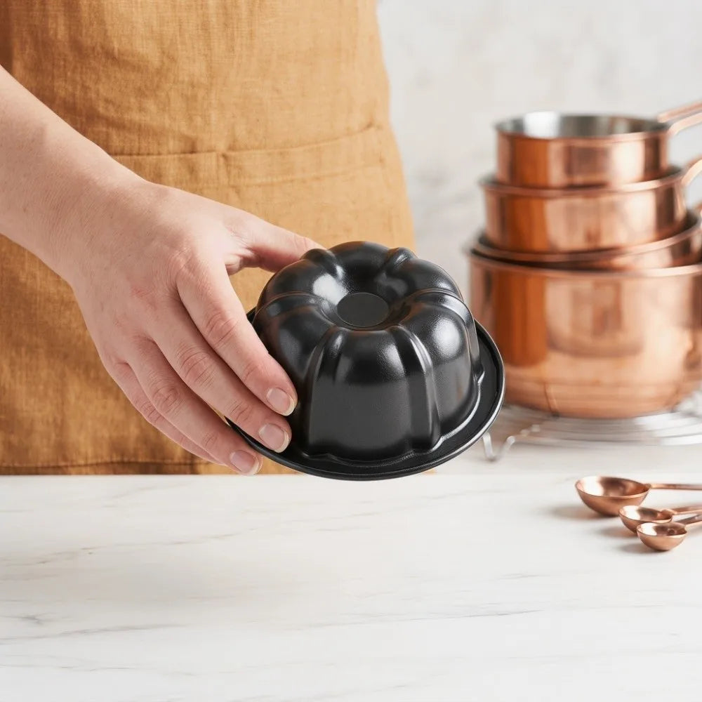A hand holds a black nonstick mini fluted cake pan above a stack of copper baking pans on a white countertop.