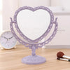 Espejo de maquillaje en forma de corazón morado con marco ornamentado sobre una superficie de madera.