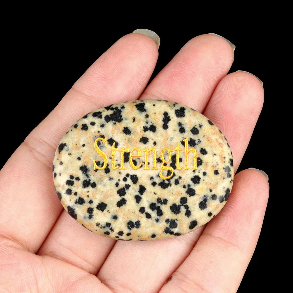 A hand holds a smooth, oval, speckled natural crystal with the word Strength inscribed on it.