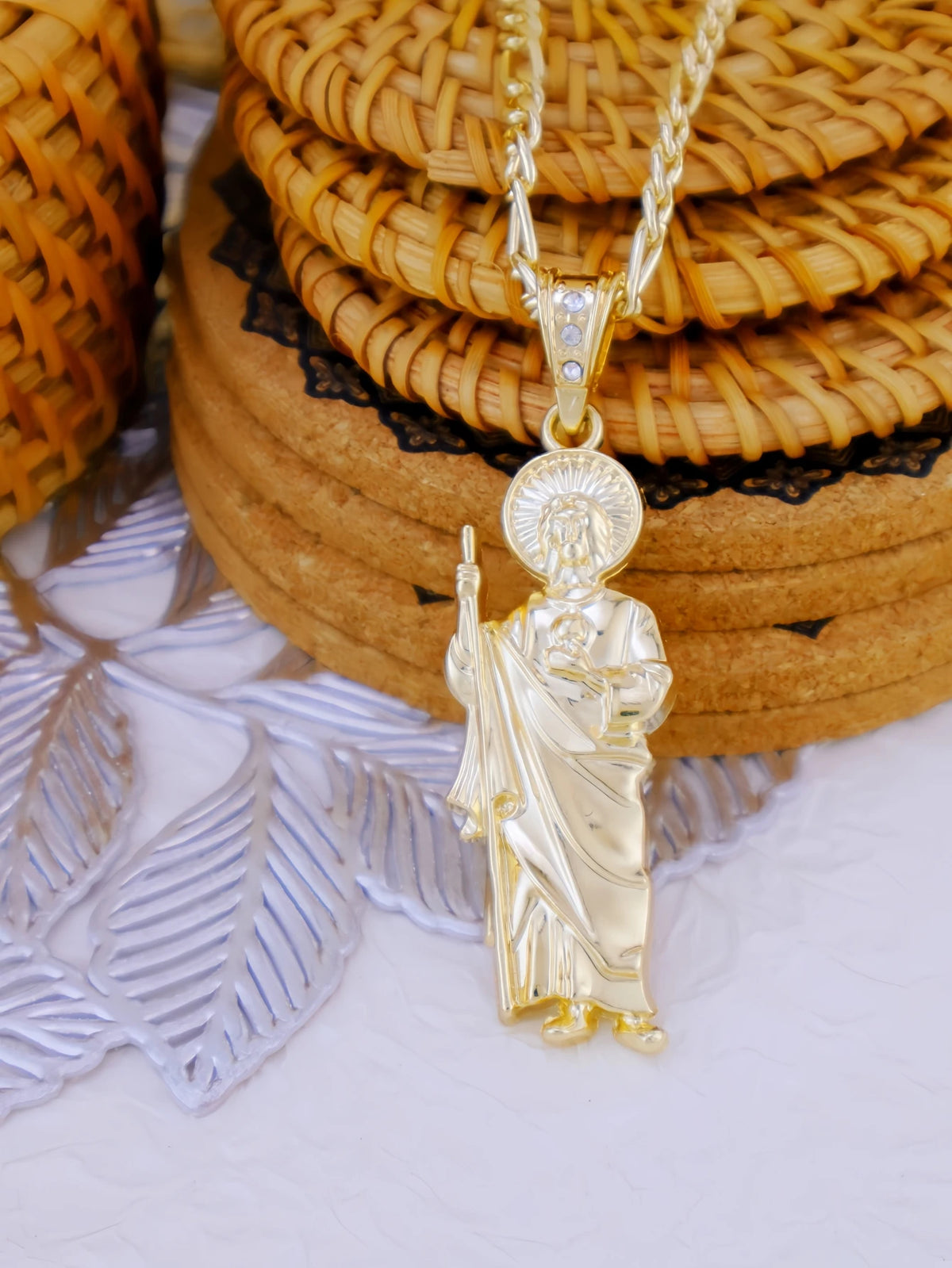 A gold-plated pendant necklace featuring a figure of Saint Jude, set against a background of woven baskets and natural wood.