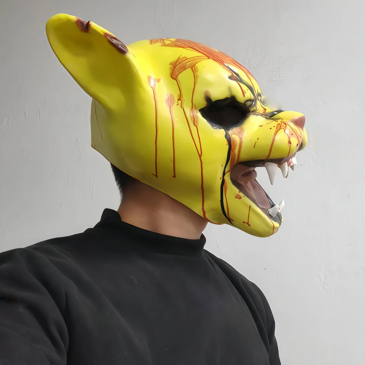 A person wearing a yellow latex mask resembling a tiger with dripping blood, against a light gray background.