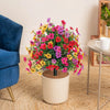 A potted artificial eucalyptus violet flower arrangement with vibrant pink, yellow, and red blooms on a light-colored stand.